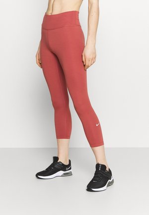 Strakke, hoog getailleerde, roodroze cropped legging gedragen met zwart-witte Nike sneakers bij een staand persoon.
