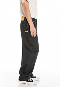Svarte denimbukser med avslappet passform, med en gul logo-patch foran og bak, sydd detaljer og sidelommer.
