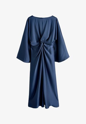 Robe en satin bleu marine avec un détail de nœud torsadé à la taille, manches longues évasées et silhouette longue jusqu'au sol. Des talons dorés à fines lanières apportent un contraste.