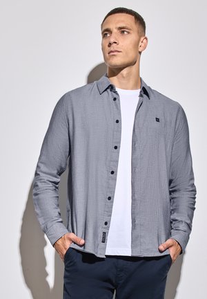 Camisa gris de manga larga con tejido texturizado, cierre de botones en la parte delantera, cuello puntiagudo y un pequeño logotipo en el lado izquierdo del pecho, llevada sobre una camiseta blanca.
