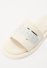 Sandale diapositive en crème avec une large sangle douce comportant un accent argenté réfléchissant et un logo en relief. Texture lisse et forme arrondie.
