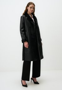 Manteau en cuir noir avec un design à double boutonnage, col en fausse fourrure et poignets. Texture lisse, longueur jusqu'au genou, associé à un pantalon noir et des talons.