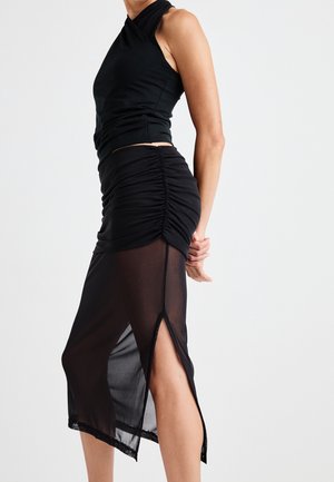 Černý halter top a nařasená černá sukně s průhledným přehozením a bočními rozparky. Materiál je hladký, což vytváří přiléhavou siluetu.