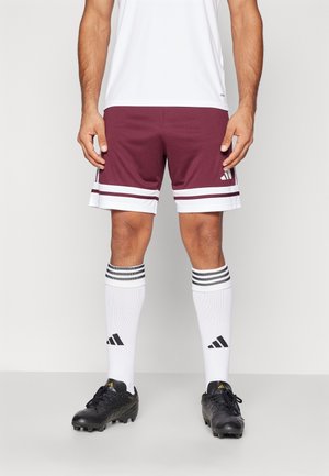 Man wearing maroon and white sports shorts, white knee-high Adidas socks with black stripes, and black cleats against plain background.