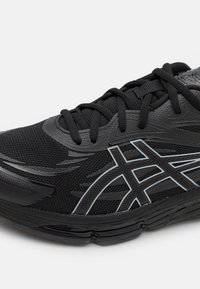 Chaussure de sport noire avec un dessus texturé, une semelle extérieure en caoutchouc et des accents réfléchissants. Les lacets et les détails structurés améliorent le soutien.