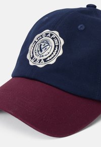 Marineblå kasket med en kastanjebrun skygge, med et cirkulært broderet logo med hvide detaljer. Stoffet har en tekstureret overflade.