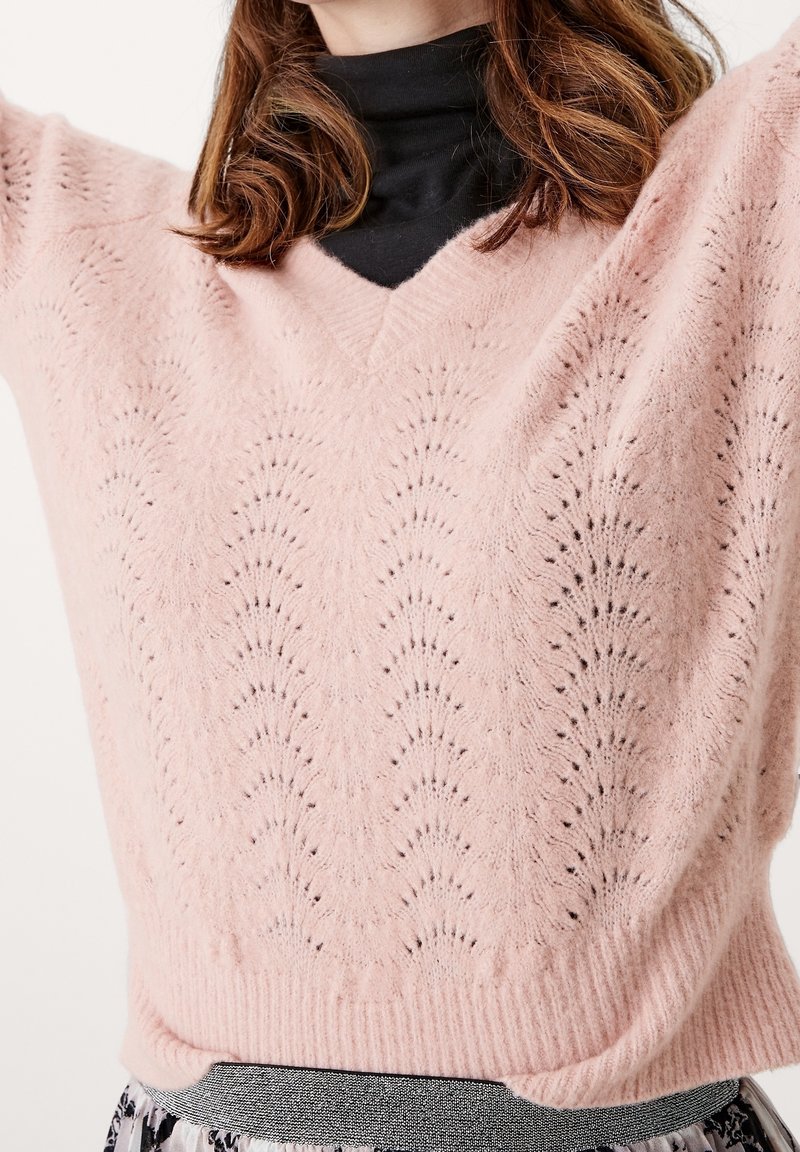 Femme portant un pull en maille rose pâle avec un motif festonné sur un col roulé noir, bras levés, et une jupe à motifs visible à la taille.