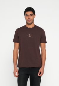 Καφέ βαμβακερό T-shirt με κοντά μανίκια, στρογγυλή λαιμόκοψη και λογότυπο με τα γράμματα "CK" σε γκρι χρώμα. Εφαρμόζει άνετα πάνω στο σώμα.