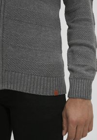 Grauer Strickpullover mit strukturierten Mustern und gerippten Bündchen. Enthält ein kleines braunes Markenetikett auf der linken unteren Seite.