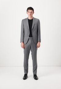 Grauer, taillierter Anzugjackett mit Revers, passende, schmal zulaufende Hose und einem schwarzen Rundhals-Shirt. Das Modell steht vor einem weißen Hintergrund.