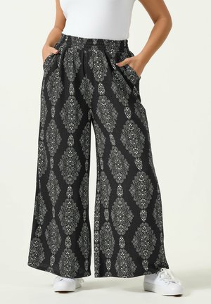 Frau trägt schwarze Hosen mit weitem Bein und weißen, filigranen geometrischen Mustern, seitlichen Taschen, kombiniert mit weißen Sneakern und einem weißen ärmellosen Oberteil.