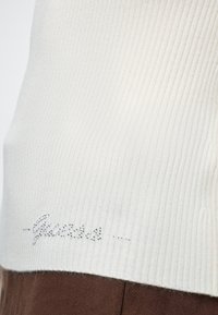 Gros plan de tissu blanc côtelé avec de petits strass formant le mot "Guess" près de l’ourlet sur un vêtement marron.