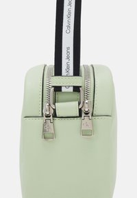 Jasnozielona skórzana torba z podwójnymi srebrnymi suwakami, kontrastującym czarnym paskiem z napisem "Calvin Klein Jeans", zaokrąglony kształt, gładka faktura.