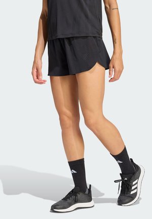 Osoba ubrana w czarne sportowe krótkie spodenki, czarne skarpetki za kostkę i czarne buty do biegania Adidas stojąca na jasnym tle.