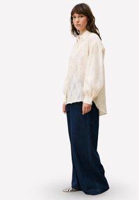 Licht crèmekleurige, gestructureerde blouse met een knoopsluiting aan de voorkant en ballonmouwen; gecombineerd met donkerblauwe, brede ribfluwelen broek en zilveren schoenen.