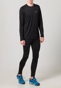 Svart långärmad träningsskjorta i fukttransporterande material, åtsittande svarta leggings och blå löparskor med ett mönstrat mönster.