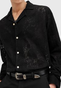 Camicetta nera a maniche corte con un motivo testurizzato sottile, dotata di colletto e bottoni beige, abbinata a una cintura nera con fibbia argentata.