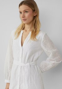 Vestito bianco a maniche lunghe con ricamo ad occhiello e chiusura frontale con bottoni. Include una cintura coordinata e una vestibilità morbida.