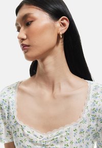 Blus med blommönster i vitt med blå blommor. Har en fyrkantig halsringning med spetsdetalj, korta ärmar och en rynkad detalj fram.