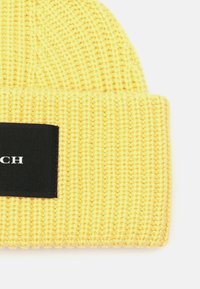 Gorro de punto amarillo con textura acanalada y puño doblado, que presenta un parche negro con letras blancas.
