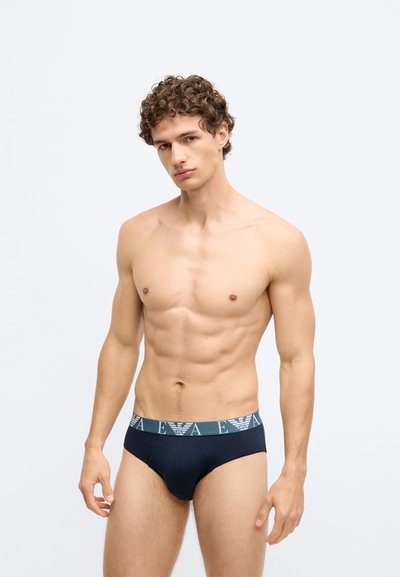 Cuecas pretas com uma cintura em turquesa que apresenta um logótipo. O tecido parece liso, com um design ajustado e contornado e costuras mínimas.