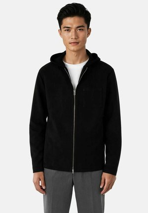 Ung mann iført en svart hettejakke med glidelås over en hvit skjorte og grå bukser, stående mot en ensfarget lys bakgrunn.