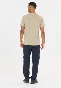 Homme debout, de dos, portant un T-shirt beige à manches courtes, un pantalon convertible bleu marine et des chaussures de sport noires sur fond blanc.