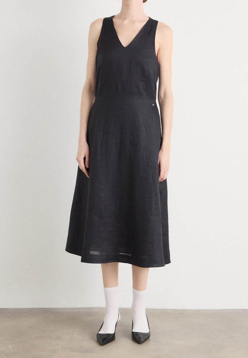 Femme portant une robe midi noire sans manches avec un col en V, des chaussettes blanches aux chevilles et des talons pointus noirs, debout sur un sol gris contre un mur blanc.