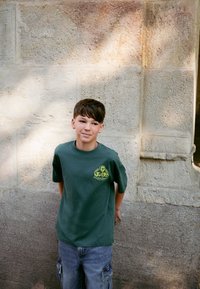 Camiseta de algodón verde con un diseño gráfico amarillo, combinada con pantalones cortos cargo de mezclilla. El fondo presenta elementos de pared de piedra con textura.