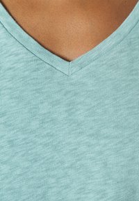 Detailní záběr na náhled s opálenou pletí, oblečenou v světle modrém pleteném tričku s V-výstřihem.