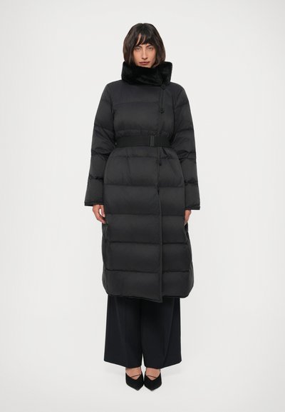 Vrouw in een lange zwarte puffermantel met een hoge bontkraag, een ceintuur op de taille, brede zwarte broekspijpen en zwarte puntige schoenen.