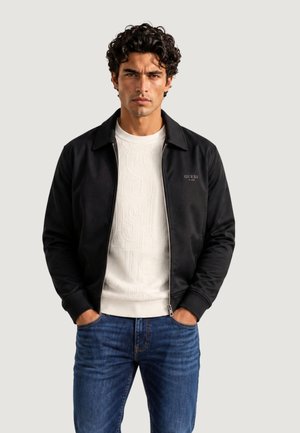 Homme aux cheveux bouclés et foncés portant une veste zippée noire par-dessus un pull blanc texturé et un jean bleu, mains dans les poches, fond neutre.