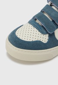 Modrá semišová teniska s krémovou perforovanou kůží a modrými akcenty. Má suché zipy a texturovanou gumovou podrážku pro lepší přilnavost.