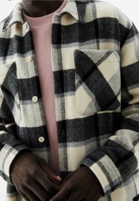 Man in een zwart, wit en grijs geruite button-up jas over een mauve sweatshirt, met handen zachtjes samengevoegd op taillehoogte.