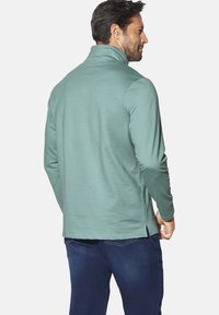 Mint green long-sleeve pullover with a high collar, side slits at the hem, and a smooth texture. Worn over dark denim pants.