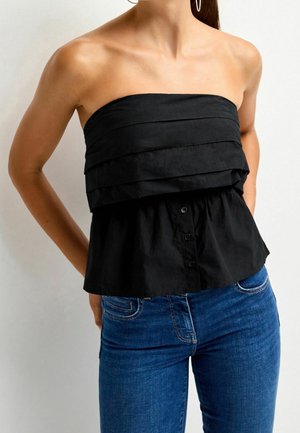 Femme portant un haut noir sans bretelles à couches et un jean bleu, les mains dans le dos, sur un fond clair uni.