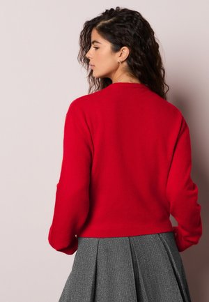 Kohver juustega naine, kes kannab punast kampsunit ja halli voltidega seelikut, seisab vasakule pööratud kehahoiakus ühetaustalise hele tausta ees.