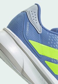 Blauwe sportieve schoen met een gestructureerd mesh bovenmateriaal, grijze accenten, een opvallende gele streep en een witte tussenzool met een lichtgewicht ontwerp.