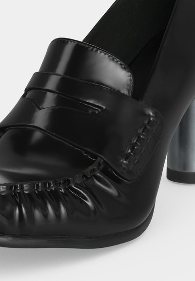 Gros plan d'une chaussure à talon haut pour femme en cuir noir brillant, avec un bout arrondi, des détails froncés et un large talon bloc métallique.