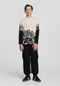 Beżowa koszulka z czarnym nadrukiem graffiti, nałożona na czarną bluzę z długim rękawem. Połączona z czarnymi spodniami typu cargo i czarnymi trampkami z białymi akcentami.