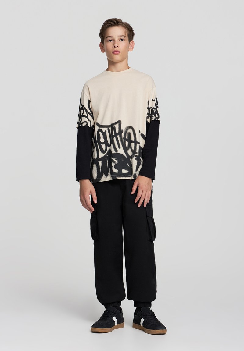 Beżowa koszulka z czarnym nadrukiem graffiti, nałożona na czarną bluzę z długim rękawem. Połączona z czarnymi spodniami typu cargo i czarnymi trampkami z białymi akcentami.