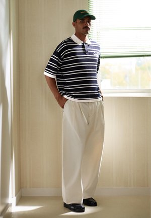 Mand står ved vindue iført grøn kasket, marineblå og hvide stribede polo, brede beige bukser og sorte sko i et blødt oplyst rum.