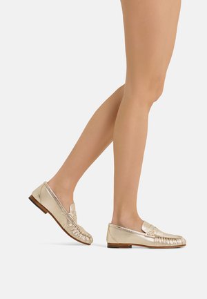 Woman's legs wearing metallic gold leather loafers with low wooden heels on a plain light background.