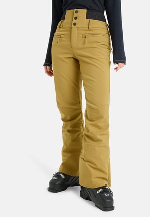 Personne portant un pantalon de ski jaune moutarde avec fermetures éclair à l'avant et fermeture à trois boutons, un haut noir à manches longues et des chaussures de ski noires.