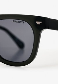 Czarne prostokątne okulary przeciwsłoneczne z ciemnymi soczewkami. Posiadają subtelny metalowy akcent na oprawie. Logo "Superdry" wytłoczone w lewym górnym rogu.