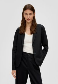 Vrouw met lang bruin haar draagt een zwarte blazer en broek over een witte tanktop, staande tegen een effen lichte achtergrond.