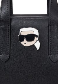 Borsa a tracolla nera in pelle con una spilla in stile cartone animato raffigurante una persona con capelli bianchi e occhiali da sole neri, attaccata sul davanti.