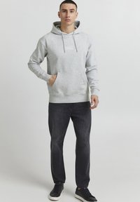 Hoodie grigio chiaro con tasca frontale, cappuccio con cordino e dettagli logo sottili. Indossato con jeans scuri e sneaker nere.