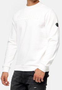 Hvid sweatshirt med et tekstureret logo på fronten, ribbede manchetter og kant, samt et sort logopatch på venstre ærme. Bæres med mørke jeans.