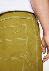 Lähikuva henkilöstä, jolla on päällään oliivinvihreät housut, joissa on valkoiset tikkaukset ja kirjailtu Nike-logon swoosh takataskussa, käsi lepäämään sivulla.